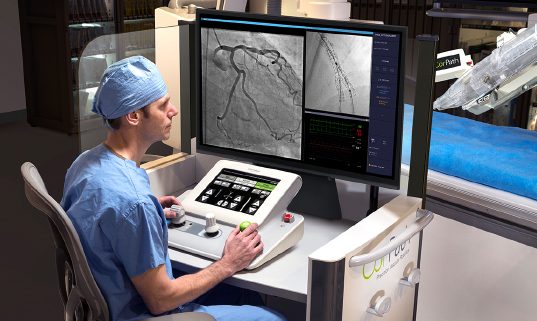 This image shows a physician seated at the interventional cockpit. The monitor in the cockpit shows a video of the stent procedure. The robotic arm is in the background of the image.