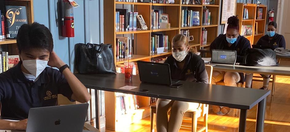 四名学生，一男三女，在图书馆学习。每个人都坐在自己的办公桌前，相距6英尺，戴着口罩，用笔记本电脑工作。