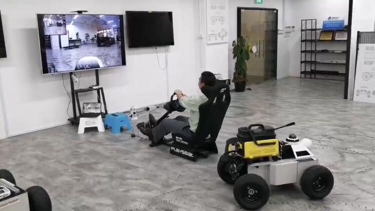 机器人显示在前景中。一名操作员坐在显示机器人周围环境的大屏幕前。操作员正在用方向盘控制机器人。